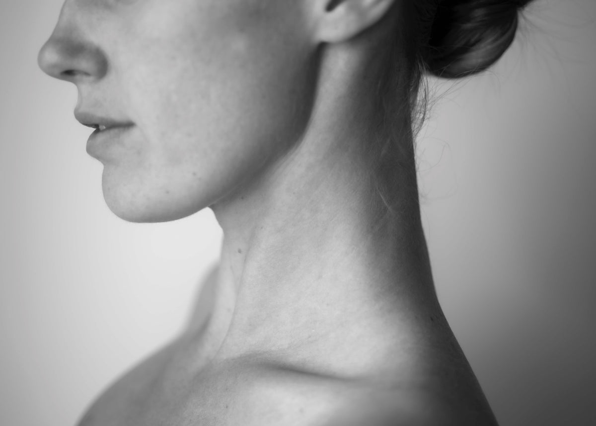 Black and white close-up profile of a woman's neck, jawline, and upper shoulder against a soft gray background. Skin Studio