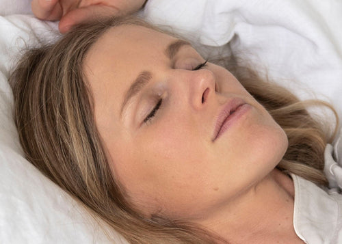 A woman lying in bed under white sheets and pillows, wearing light beige pajamas. A small wooden bedside table with a lamp is visible in the background.