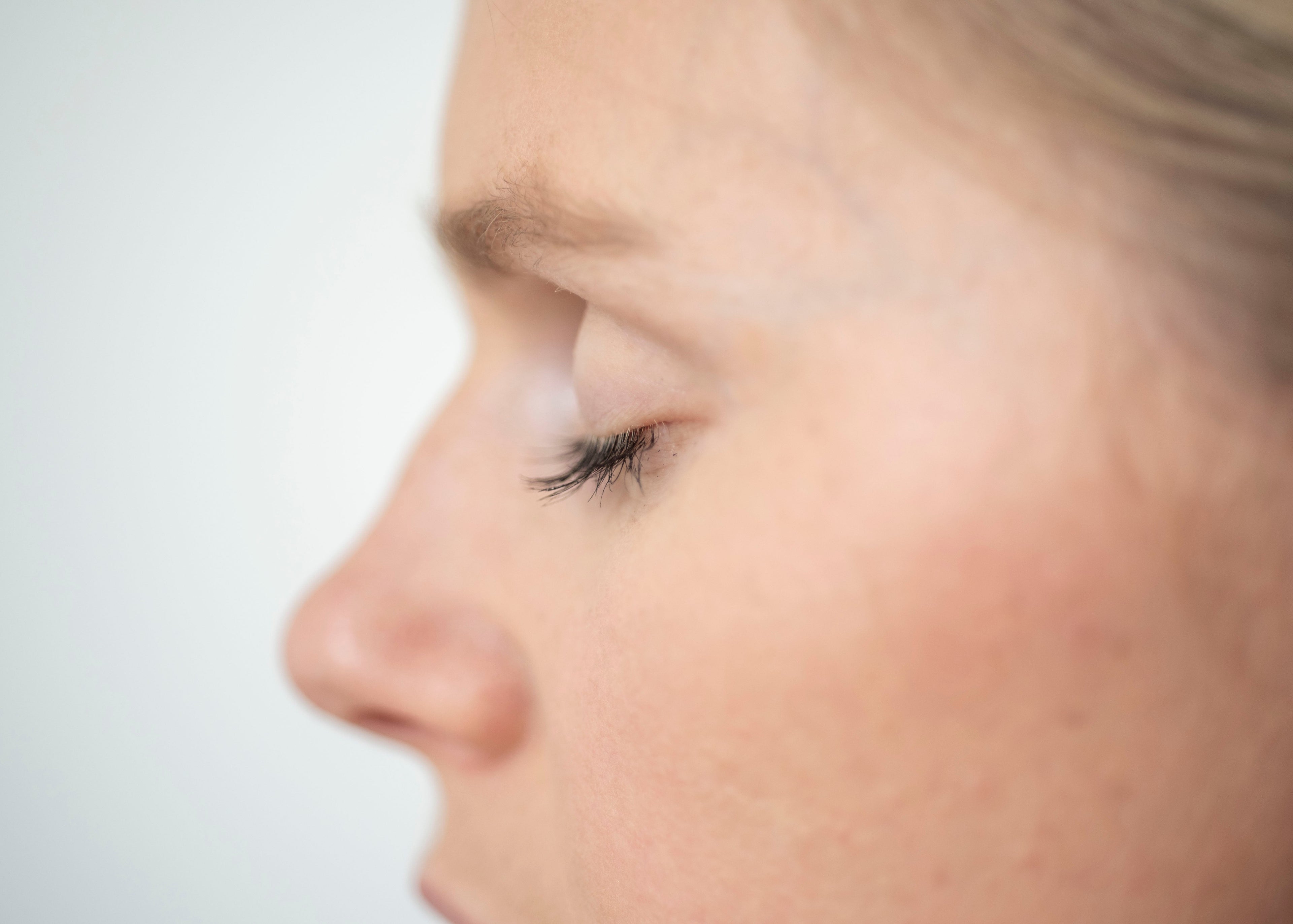 Close-up side profile of a woman's face showing her eye, nose, cheek, and part of her hair against a light gray background.