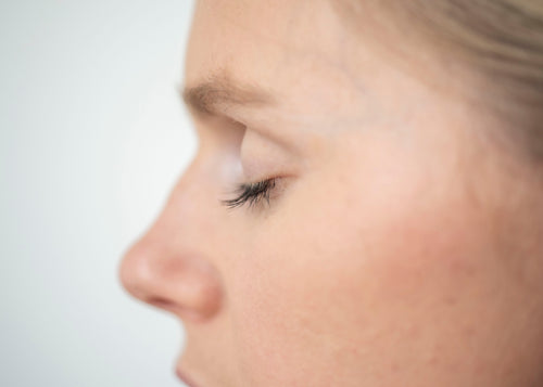 Close-up side profile of a woman's face showing her eye, nose, cheek, and part of her hair against a light gray background.