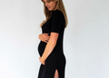 A pregnant woman wearing a fitted black short-sleeve maternity dress with a thigh-high slit, standing in profile against a plain light gray studio background.