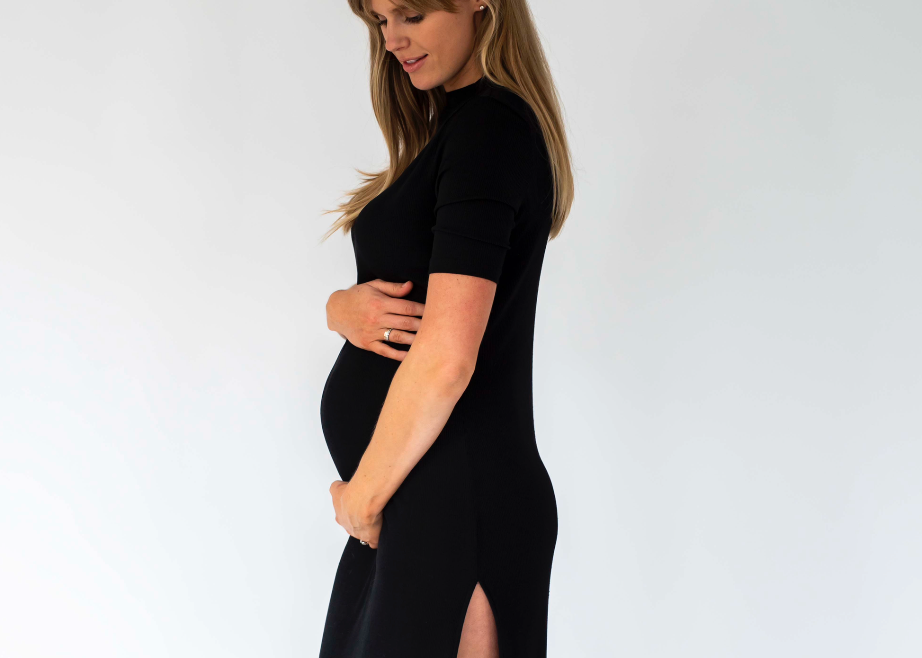 A pregnant woman wearing a fitted black short-sleeve maternity dress with a thigh-high slit, standing in profile against a plain light gray studio background.